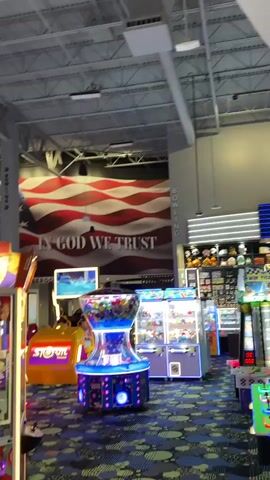 The video captures the interior of a spacious, modern entertainment complex, likely an arcade and bowling alley, with high ceilings and exposed ductwork. The camera pans across various arcade games, including claw machines and other brightly lit cabinets, featuring text like "STORM" and "OCITY HOUSES."

A prominent feature is a large wall mural depicting the American flag with the phrase "IN GOD WE TRUST" written across it. Nearby, a sign for "BOWLING" is visible. The complex appears to be multi-level, with an upper floor or mezzanine area visible, marked by signs for "SWEETROOMS" (possibly "RESTROOMS") and "ROOMS."

A unique decoration is a white race car, reminiscent of an IndyCar, mounted vertically on a wall, with "TEXACO" and "1" visible on its side. Below this, a "DINING" sign is partially seen. The facility features a distinctive patterned carpet and colorful wall panels in blue, green, and yellow. As the video concludes, a woman walks by in the foreground. The overall ambiance suggests a lively, well-lit recreational venue.