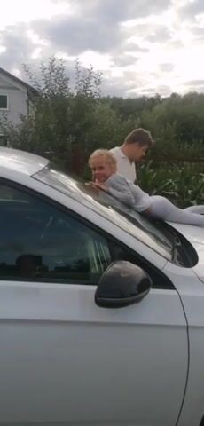 This video captures a lighthearted moment outdoors, likely in a residential or semi-rural area given the presence of a house and greenery. A young girl with blonde hair is playfully lying face down on the hood of a white car, her body stretched out. She is wearing a grey long-sleeved shirt and light-colored pants, with pink shoes. Her expression is a playful smile directed towards the camera.

Behind her, a man in a light-colored polo shirt is standing next to the car, his attention seemingly focused on the girl. Another man, partially visible in the foreground with his back to the camera, is also present. The scene appears to be set during the late afternoon or early evening, with soft, overcast lighting, suggesting cloudy weather. The backdrop consists of trees and bushes, with a wooden fence visible in the distance. The overall mood is casual and familial. No text is visible in the video.