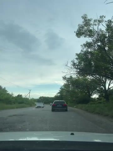 The video shows a view from inside a car driving on a paved road. The road is partially damaged with potholes. Several cars are driving ahead on the same road. The surroundings are covered with lush green trees and vegetation on both sides of the road. A few utility poles are visible on the left side of the road. The sky is overcast with grey clouds. The camera seems to be mounted on the dashboard of the car.