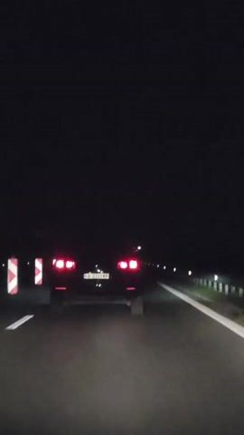 The video shows a rear view of a black car driving on a highway at night. The car's red taillights are illuminated. The road is paved and has a white line marking the lane. On the left side of the road, there are several reflective traffic cones or markers with red and white diagonal stripes. In the distance, there are streetlights or other illuminated markers along the side of the road. The sky is dark, indicating it is nighttime. The camera appears to be mounted on another vehicle following the black car.