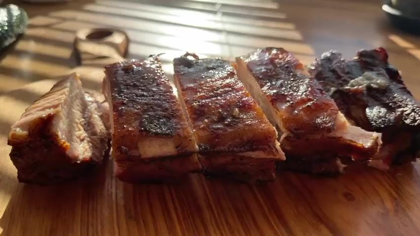A close-up, medium shot focuses on sliced pork ribs presented on a wooden cutting board. The ribs, glazed and glistening, are arranged in a row, with some sections displaying a dark, caramelized crust and others showing the tender, pink meat beneath. The lighting suggests it is daytime, with sunlight casting shadows from a blind onto the background, creating a striped pattern. The overall impression is one of a prepared meal, likely a barbecue or roasted dish. The background is slightly out of focus, but hints at a wooden surface and possibly a small decorative item.