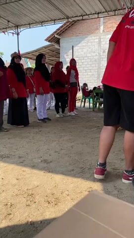The video shows a group of people, mostly women wearing red shirts and headscarves, standing in a line outdoors. They appear to be participating in some sort of event or activity. The ground is dirt and there is a makeshift roof overhead, supported by poles. In the background, there are buildings and a few trees. One person in the foreground, wearing a red shirt and black shorts, is facing away from the camera, obscuring the view of the people in the line. The lighting suggests it is daytime, likely sunny. The people in the line are looking in the same direction, towards the person filming or someone in front of them. Some of the women are wearing white pants, while others are in black pants. One woman is wearing a beige hat. The overall impression is of an organized gathering or a warm-up for an event.