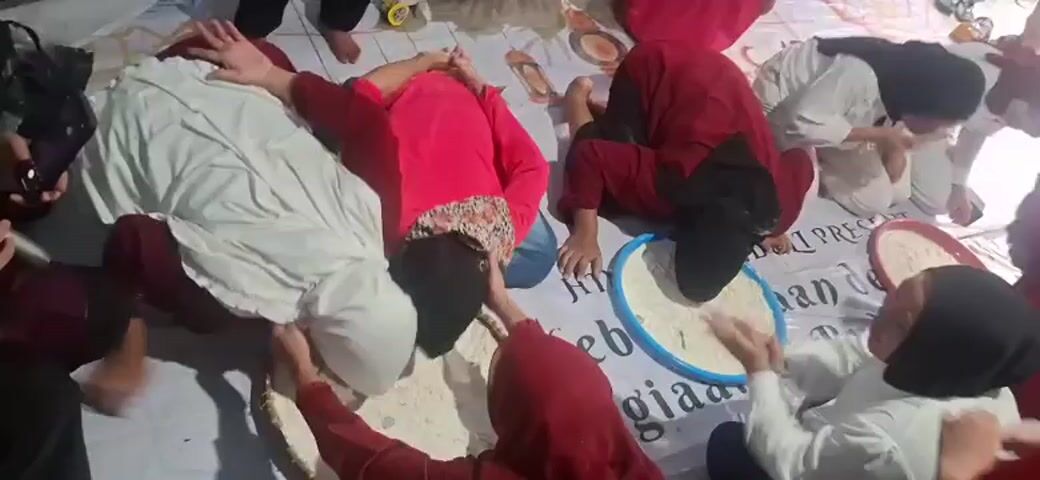 A group of people, mostly women and children, are gathered on the floor, which is covered by a white tarp with some text printed on it. They are in a low-lit room with tiled walls. Some individuals are wearing hijabs. The people appear to be participating in a game or activity involving flour. Several large, flat woven mats and a round blue tray are spread on the tarp, filled with a white powdery substance that looks like flour. Some of the participants are leaning over these containers, and it appears they are trying to eat or move the flour with their mouths or hands. One person is holding a microphone. Some individuals have flour on their faces and clothes. The overall atmosphere seems to be one of playful activity, possibly a competition or celebration.