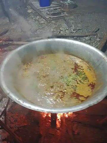 The scene is a cooking setup outdoors with a large metal pot, probably aluminum, sitting over an open wood fire. The pot contains a bubbling stew or soup that is releasing steam. The stew is a mixture of colors, including yellow, red, and green, suggesting a variety of ingredients. The fire is burning brightly under the pot, providing the heat for cooking. To the left of the pot is a rough, stone or concrete structure. There are also some miscellaneous items like a blue container and possibly some discarded packaging near the stone structure. The overall setting suggests a rustic or informal cooking environment.