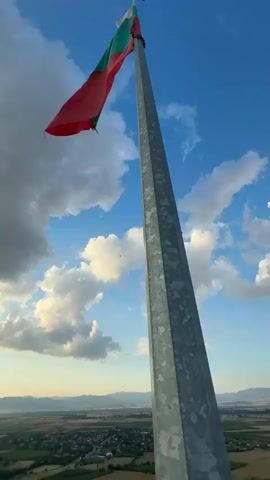 This is a video capturing a panoramic view from a hilltop in Panaretovtsi, Bulgaria. The primary subjects are the Bulgarian flag flying proudly from a tall metal flagpole and the expansive landscape below. The scene depicts a rural setting with rolling hills, agricultural fields, and a distant town nestled in a valley.

The activity appears to be a gentle panning movement, starting with a close-up of the flag against a bright blue sky dotted with fluffy white clouds, then transitioning to a wider view of the countryside. The camera then shifts focus to capture the setting sun casting a warm glow over the horizon, creating a sense of depth and tranquility. The time of day is clearly sunset, evidenced by the golden light and long shadows. The weather appears pleasant, with a mix of sun and clouds contributing to a dramatic sky.

Notable details include the vibrant colors of the flag, the textured surface of the flagpole, and the patchwork of green and brown fields below, suggesting a thriving agricultural area. The distant mountains add a sense of scale and natural beauty to the location. The overall mood conveyed is one of peace, national pride, and appreciation for nature's beauty. There is no visible text in the video.