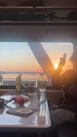 The video shows a person sitting at a table by a window on a train. The sun is setting outside, casting a warm glow on the scene. On the table are a water bottle, a can of soda, a cup, an apple, and a notebook. The person is holding a phone up to their face, possibly taking a picture or video of the sunset. The train compartment appears to be cozy and well-lit, with luggage visible on the overhead shelf. The overall atmosphere is one of relaxation and appreciation for the beautiful scenery.