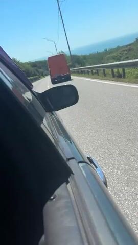 The video, shot from the passenger side of a moving car, shows a scenic drive along a coastal road under a clear blue sky. The car is overtaking an orange commercial van with a Russian license plate "M 664 CC 123." The road has a white solid line on the right shoulder and a dashed white line separating lanes, indicating a multi-lane or passing section.

To the right of the road, a metal guardrail separates the asphalt from a downward slope covered in green vegetation. In the distance, a large body of water, likely the sea, is visible, extending to the horizon. Trees and green hills line the landscape between the road and the sea. Street lights or power poles are visible along the road, suggesting infrastructure. Other cars can be seen in the far distance ahead on the road. The weather is bright and sunny, indicative of daytime.