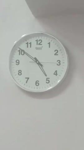 THE VIDEO SHOWS A CLOSE-UP OF A ROUND WHITE WALL CLOCK. THE CLOCK HAS BLACK NUMBERS INDICATING THE HOURS AND SMALL BLACK DASHES INDICATING THE MINUTES. THERE ARE TWO BLACK HANDS ON THE CLOCK. THE HOUR HAND IS POINTING SLIGHTLY PAST THE NUMBER 10. THE MINUTE HAND IS POINTING SLIGHTLY PAST THE NUMBER 5. A BRAND NAME, "Ajanta", IS VISIBLE NEAR THE NUMBER 12. THE CLOCK IS MOUNTED ON A PLAIN, LIGHT-COLORED WALL, POSSIBLY WHITE OR OFF-WHITE. THE LIGHTING IS SOFT AND EVEN, SUGGESTING AN INDOOR ENVIRONMENT. THE VIDEO IS SLIGHTLY BLURRY AND UNSTEADY, WITH MINOR SHIFTS IN FRAMING AND FOCUS THROUGHOUT THE SHORT DURATION. THE BACKGROUND CONSISTS ENTIRELY OF THE WALL AND A SMALL PORTION OF WHAT APPEARS TO BE A CEILING OR CORNER AT THE TOP LEFT OF THE FRAME. NO PEOPLE OR OTHER OBJECTS ARE VISIBLE. THE FOOTAGE IS IN COLOR.
