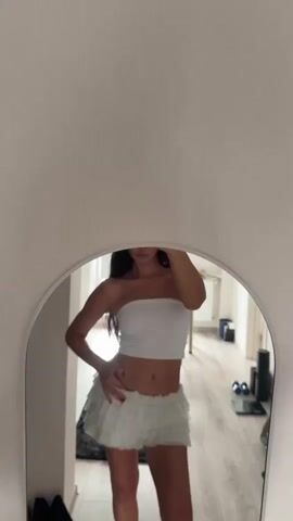 A woman stands in front of a mirror, taking a selfie with her phone. She is wearing a white tube top and a white ruffled skirt. Her hair is dark and falls past her shoulders. Her right hand is on her hip, and her left hand holds her phone up to take the picture. The mirror reflects a room with light-colored walls and a wooden floor. There are some objects visible in the background, including a rug, shoes, and what appears to be gym equipment. The lighting is bright, and the overall impression is of a home environment.