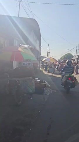 A street market scene unfolds. A vendor's cart with a colorful umbrella displays "SALAK PONDOH 6.000" signs, alongside piles of vegetables. Various individuals are seen on motorbikes and walking through the bustling market. Stalls line both sides of the street, with goods and produce on display. The sky is clear and blue, with sunlight casting shadows.