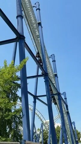 This is a low-angle video of a roller coaster climbing a steep track against a clear blue sky. The roller coaster train, with several passengers visible, is being pulled upwards by a chain lift mechanism. The track and its supporting structure are painted in shades of blue and white. Lush green trees are visible on the left side of the frame, suggesting an outdoor amusement park setting. The bright sky and the presence of leaves indicate it is daytime, likely during fair weather. The scene evokes anticipation and excitement, common emotions associated with roller coaster rides. There is no visible text in the video. The location is Castelnuovo del Garda, Italy.