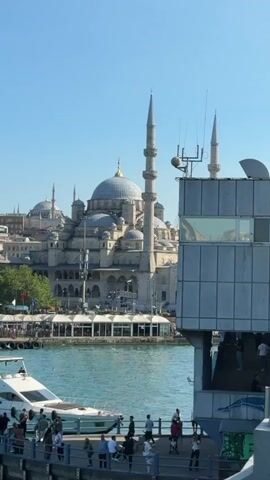 The video shows a view across a body of water, likely a harbor or bay, towards a large, ornate mosque with multiple domes and tall minarets. In the foreground, a concrete structure with windows juts into the frame from the right, and a portion of a white boat is visible on the left. A walkway or pier runs along the edge of the water in the foreground, with several people standing and walking on it. The water is a vibrant blue-green, and the sky is clear and blue. The mosque appears to be made of light-colored stone and is intricately detailed. A few birds are visible in the sky.