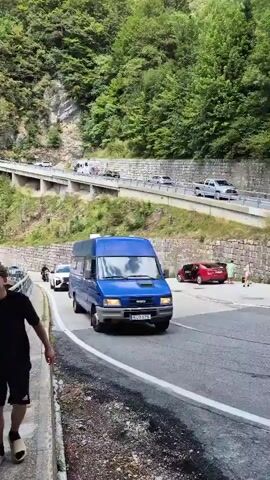 A blue van is driving on a winding mountain road. Cars and motorcycles are also on the road, as well as people walking along the side of the road. The road is lined with a metal guardrail and a stone wall. The mountain is covered in trees. The sky is cloudy.