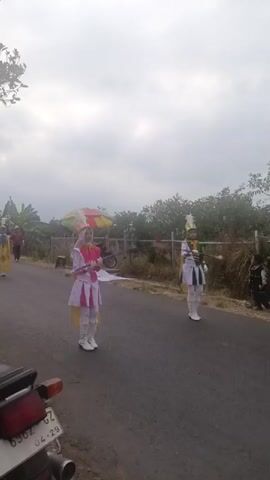 Two individuals in marching band uniforms stand on an asphalt road. The person on the left wears a white and pink uniform with a hijab, holding a baton. The person on the right wears a white and green uniform and also holds a baton. In the background, a person in a yellow outfit walks away from the camera, and other figures are visible further down the road. Trees and foliage line the side of the road, and the sky is overcast. A motorcycle is partially visible in the foreground on the left. The individuals appear to be participating in a parade or performance.