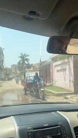 The video, filmed from inside a car in Nasheman Colony, Pakistan, shows a dusty, unpaved residential street on a sunny day. The camera looks through the windshield as the car moves forward.

In the rearview mirror, the driver, a man with a beard, is visible, looking focused.

Initially, a man on a motorcycle attached to a red trailer carrying multiple blue water bottles is seen in front, then passes by. Further down the street, two other men on motorcycles are visible, and one walks in the distance. The street is lined with houses, some with walls and fences, and a prominent palm tree stands in the background. Power lines are also visible overhead.

The dashboard of the car is visible at the bottom of the frame, with the text "clarion" on what appears to be the car's audio system. The sky is clear, indicating bright daylight.
