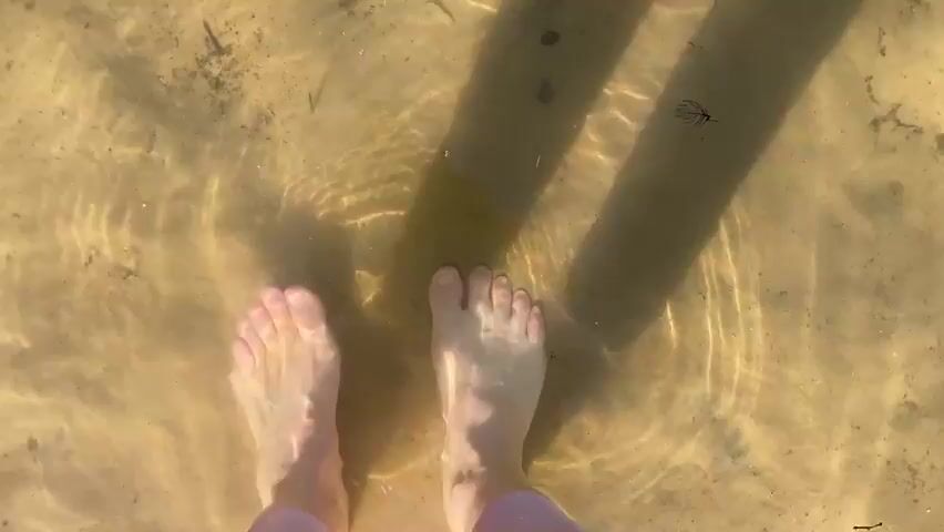 This is a top-down shot of a person's bare feet standing in shallow, clear water. The water is rippling, creating patterns of light on the sandy bottom. Two dark shadows of what appear to be legs are cast on the water's surface, running parallel to each other. There are small particles of dirt and debris visible on the bottom and floating in the water. The feet are pale and appear to be moving slightly, disturbing the sand beneath them. The overall impression is one of a peaceful, natural setting, perhaps a lake or a shallow stream.