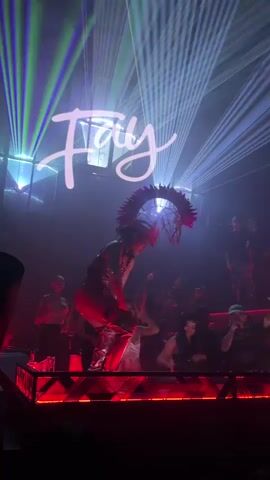 A person in a revealing outfit and elaborate headdress performs on a stage in a dimly lit club. Laser lights beam down from above, creating a vibrant atmosphere. Several people are visible in the background, some dancing and others watching the performance. The stage is illuminated with red lights. The word "Fray" is displayed in white script across the top of the frame.
