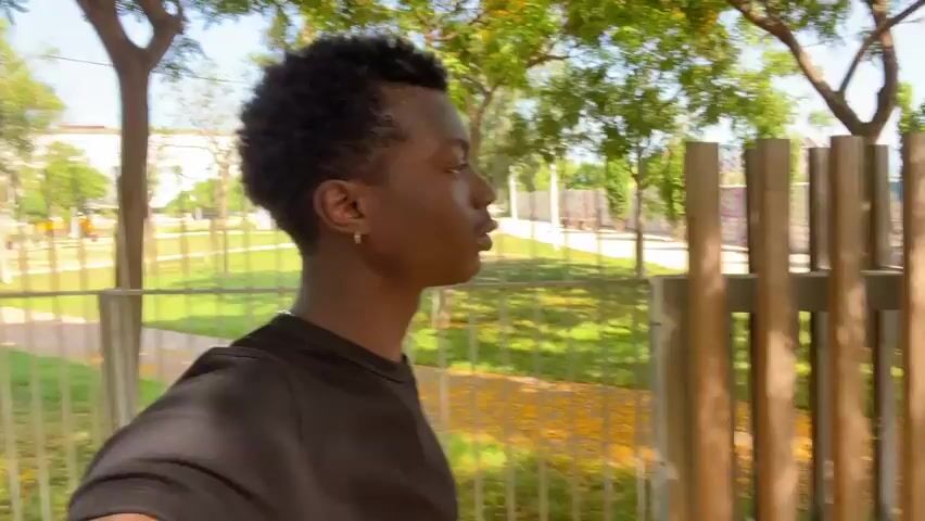 This media file features a young Black man walking in an outdoor park. He is wearing a black t-shirt and has short, curly dark hair. A small hoop earring is visible in his left ear. The man is looking off to the side, with his profile visible for most of the clip, before turning his head to look directly at the camera.

The setting appears to be a park or public space with a walking path, a metal fence in the background, and a wooden fence or structure in the foreground. Trees with green and yellow foliage are present, suggesting it might be late spring or summer. The sunlight is bright, indicating it is daytime, likely late morning or afternoon. The overall mood seems relaxed and casual. There is no visible text in the media file. The location context places this scene in Sant Adrià de Besòs, Spain.