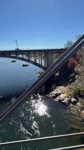 The video shows a clear blue sky above a river spanned by a concrete bridge. Several people on paddleboards and kayaks are visible in the water below the bridge. A white truck is driving across the bridge, and there are rocks and trees on the riverbanks. The sun is shining brightly, reflecting off the water's surface. The foreground features a metal railing.