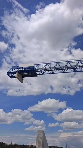 A blue construction crane is shown against a cloudy blue sky. The camera pans up, focusing on the crane's operator cabin and boom. The cabin is white with glass windows, and a person can be seen inside. The boom is a long, triangular structure. Below the crane, a blurry landscape of trees and buildings is visible.