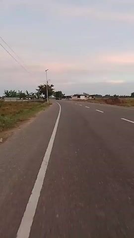 The video shows a first-person perspective of someone riding on a road. The road is paved and has a white line dividing the lanes. The ride appears to be taking place in a rural area, with grassy fields and some trees on the left side of the road. There are also some buildings visible in the distance. The sky is cloudy, and it appears to be either sunrise or sunset. The camera is moving forward, indicating the person is in motion.