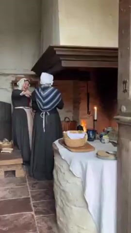 Two women dressed in historical costumes stand in a rustic kitchen. They both wear white head coverings and long, dark dresses with aprons. One woman faces the viewer, adjusting her apron, while the other has her back to the viewer, wearing a blue and white shawl. The kitchen features a large stone fireplace with a fire burning inside. A table covered with a white cloth holds bread, mugs, and other items. The room has a stone floor and is dimly lit, with a candle providing additional light. The camera is stationary and views the scene from an open doorway.
