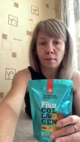 A woman sits at a table and holds a bag of fish collagen powder. She lifts a glass containing a yellow liquid to her lips and drinks it. The background is a wall with framed pictures of coffee-related items.