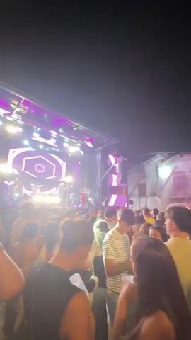 The video shows a crowded outdoor concert at night. A band is performing on a brightly lit stage with a large screen displaying a geometric pattern. The audience is densely packed, with many people holding drinks and looking towards the stage. The lighting is dynamic, with spotlights and colorful LEDs illuminating the scene. The overall atmosphere appears lively and energetic.