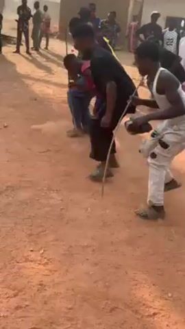 The video depicts a group of people dancing in a dusty, outdoor setting, seemingly a village or rural area.  Several individuals, mainly young men, are the focus, moving their bodies in a rhythmic way, some while wielding sticks.  They wear casual clothing, including tank tops, t-shirts, and jeans.  The ground is reddish-brown and dusty, with a light haze in the air.  Around the periphery of the dancing group, a crowd watches.  In the background are simple, low-lying structures, likely homes, and some trees. The video is handheld and shaky.