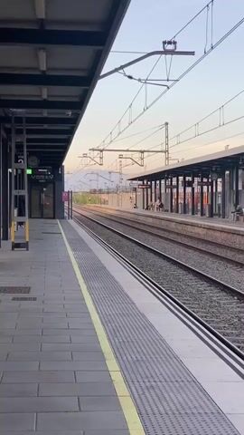 The video opens with a view of a train station platform in la Llagosta, Spain. The camera pans from left to right, showcasing two sets of railway tracks stretching into the distance. The platform is paved with grey tiles and features a yellow safety line along its edge. Overhead power lines crisscross above the tracks. On the opposite platform, a covered waiting area is visible, with several people sitting on benches. As the camera continues to pan, a fence separates the tracks from what appears to be an industrial area with cargo containers. A tall, modern-looking light tower stands between the tracks and distant hills. The camera then tilts upwards, revealing a clear blue sky where a distinct moon is visible. The scene suggests late afternoon or early evening.
