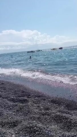 A wide-angle shot captures a pebble beach with the ocean stretching out before it. The water is choppy, with waves lapping at the shore. Several people are visible in the water, swimming and enjoying the day. In the distance, a few boats are anchored. The sky is a bright blue with fluffy white clouds. On the beach, sunbathers are relaxing on lounge chairs, and children are playing with toys in the sand. A lone bird walks along the shoreline. The overall atmosphere is one of a busy, pleasant summer day at the beach.
