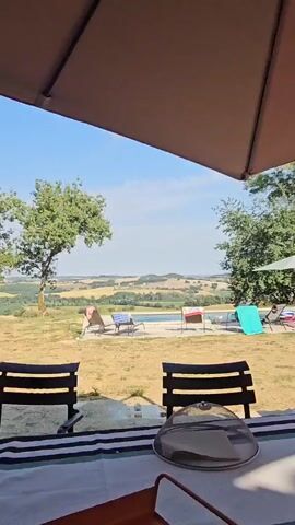The video opens with a close-up view of a table with a white and striped tablecloth. Two black chairs are visible in the foreground. A clear glass cloche sits on the table. The camera then pans out to reveal a scenic landscape with rolling hills, trees, and a swimming pool. Several lounge chairs are arranged around the pool, some with towels draped over them. The sky is clear and blue with a few scattered clouds. The overall impression is one of a peaceful, sunny day in a rural setting.