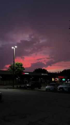 The video shows a street scene at dusk. A vibrant pink and purple sky is visible behind a row of businesses. One of the businesses has a sign that reads "bq CHICKEN". Cars are parked in front of the businesses. A street lamp illuminates the foreground. A tree with green leaves is on the left side of the frame. The overall mood is calm and serene, with the dramatic sky being the focal point.