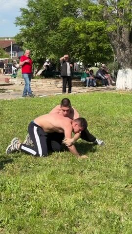 The video shows two shirtless men wrestling on a grassy lawn in a park, observed by a small crowd. They are both wearing dark pants; one has a white stripe along the side. The wrestlers are grappling on the ground. Several people are watching, including a man in a blue shirt who briefly walks across the scene in the foreground. Other observers are seated on benches in the background. There are trees in the park.