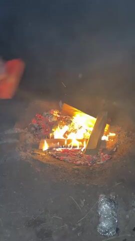 The video shows a campfire with logs and embers. There are several items wrapped in aluminum foil placed in or around the fire pit. In the background, out of focus, there are some objects that appear to be a shovel and possibly a stick or a rifle. The scene is dark, suggesting it is nighttime, and there is smoke rising from the fire.