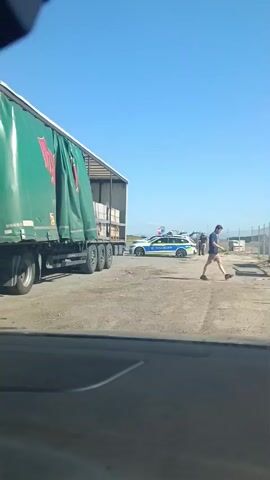 A police car with flashing blue and yellow lights is parked on a gravel surface. Several police officers are standing around the car and a large truck. One officer is in the process of opening the driver's side door of the police car. Another officer is standing near the back of the police car, and two other officers are talking to a man in civilian clothes. The truck is a large semi-trailer truck with a green tarp covering its load. The scene appears to be outdoors, with a clear blue sky overhead. The ground is covered in gravel, and there is a fence in the background.