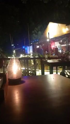 The image captures a vibrant nighttime scene at an outdoor restaurant or bar in Arambol, India. In the immediate foreground, a wooden table is illuminated by the soft, warm glow of a lit candle housed within a clear glass jar, creating an intimate ambiance. To the left of the candle, a small, dark object, possibly an ashtray or container, is visible.

Beyond the table, the establishment extends into a lively area with more wooden tables and chairs, separated by a wooden railing. Several patrons are visible, some seated and others standing, enjoying the evening ambiance. A person in a light blue shirt is prominently featured in the midground, facing away from the camera, seemingly observing the activity.

The entire scene is bathed in a mix of artificial lights. Warm string lights are draped across structures, complemented by brighter spotlights illuminating a larger, possibly covered area or stage in the distance. Some blue lighting is also visible further back, adding to the varied color palette. The setting appears to be an elevated patio or outdoor dining area, with trees and dark foliage framing the top and left edges of the frame, suggesting a natural, perhaps tropical, environment. The overall atmosphere is casual, social, and inviting, typical of a relaxed evening out in a popular tourist destination. No legible text is visible in the frame, and the clear view of the night suggests fair weather.