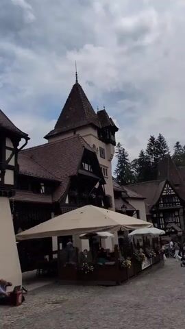 The video opens with a shot of a large building with a distinctive tower and steep, tiled roofs, reminiscent of a castle or a historical manor. The building has dark timber framing against a lighter facade. In the foreground, several large beige umbrellas provide shade over outdoor seating areas, suggesting a cafe or restaurant. People are seated at tables beneath the umbrellas, and some individuals are seen moving in the background. To the left of the frame, a person is seated on the ground, looking at a laptop placed on a red object. The ground appears to be paved with cobblestones or similar material. The sky is partly cloudy. The camera pans slightly to the left throughout the clip.