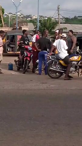 The video shows a group of people gathered in an intersection around several motorcycles and a three-wheeled vehicle. Some individuals are standing, while others are sitting on the motorcycles. A silver van is parked nearby with its driver-side door open. In the second half of the video, a woman, a child, and a man get on one of the motorcycles and drive away. The setting appears to be a street in a suburban or semi-urban area, with buildings and trees visible in the background.