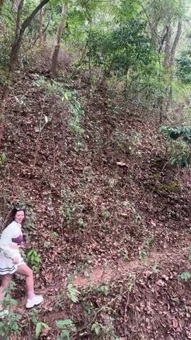 A woman in a white outfit, consisting of a short skirt, a jacket, and white sneakers, walks uphill through a forested area covered in fallen leaves.  She pauses and waves, then continues, reaching out to touch a small tree.  The forest is dense with a mix of trees and undergrowth.
