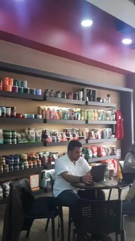 The interior of a coffee shop is shown in this video. Several people are seated at tables, including a man working on a laptop, and a couple eating. A large display shelf is stocked with bags of coffee and related merchandise. Outside the shop’s glass facade is a street with pedestrian and vehicle traffic. The ceiling is decorated with a design resembling wood grain, and a ceiling fan is visible.