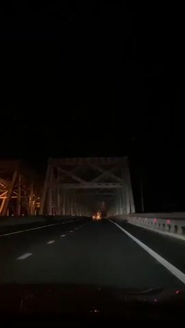 A first-person perspective video shows a car driving at night on a highway that crosses a large steel truss bridge. The car's headlights illuminate the road ahead, revealing white lane markings. The bridge structure, with its intricate metal beams and supports, looms over the highway. Distant lights from the other side of the bridge are visible in the darkness. The sky is completely black, indicating it is nighttime.