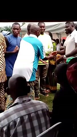 The video shows an outdoor gathering, possibly an event or ceremony, with several people interacting. In the foreground, two men are shaking hands, one wearing a blue and white patterned shirt and the other a light-colored polo shirt and pants. Behind them, other individuals are visible, some standing and some seated, under a tent-like structure. One man in a lime green long-sleeved shirt is also in the frame, and a woman in a colorful dress is visible behind him. The ground appears to be covered in grass. The overall atmosphere seems to be one of social interaction and perhaps a formal occasion.