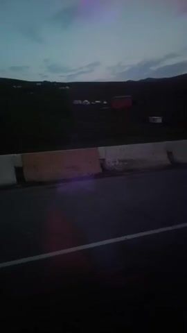 A low-angle video shows a road with a concrete barrier on the right side, and a dark landscape with some distant structures, possibly buildings and some livestock, under a twilight sky. The camera appears to be moving forward, judging by the slight blur.