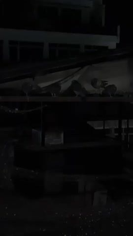 The video shows several pigeons perched on a dark structure, possibly an awning or part of a building, at nighttime. There are at least six pigeons visible, with one briefly flapping its wings around the 0:02 mark. The structure appears to be made of metal or wood, and there is some debris on its surface. In the background, a dark building with dimly lit windows can be seen. The overall scene is dark, indicating it is night. There is no visible text or discernible weather conditions. The location context provided is Karditsa, Greece.