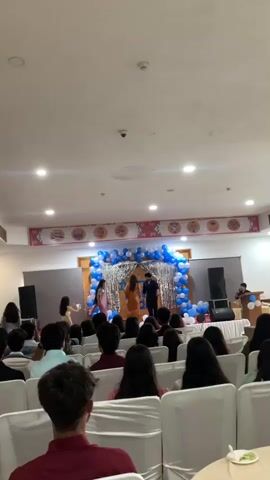 The video shows an indoor event with a stage decorated with blue and silver balloons. On stage, two individuals, a male and a female, are interacting. A banner above the stage reads "INSTITUTE NOVUS 2023". A crowd of people is seated in chairs facing the stage, with some individuals visible in the foreground from the back. Several speakers are positioned on either side of the stage. The lighting appears to be from overhead fixtures.
