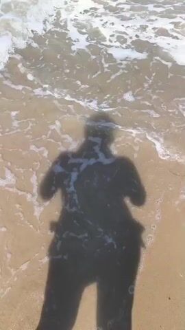 The video shows a shadow of a person on a sandy beach as waves wash over it. The camera is pointed down, capturing the shadow from above. The shadow is of a person standing with their arms slightly bent. As the video progresses, the waves cover more of the shadow, obscuring parts of it with foam and water. The environment appears to be a beach with the ocean in the background.