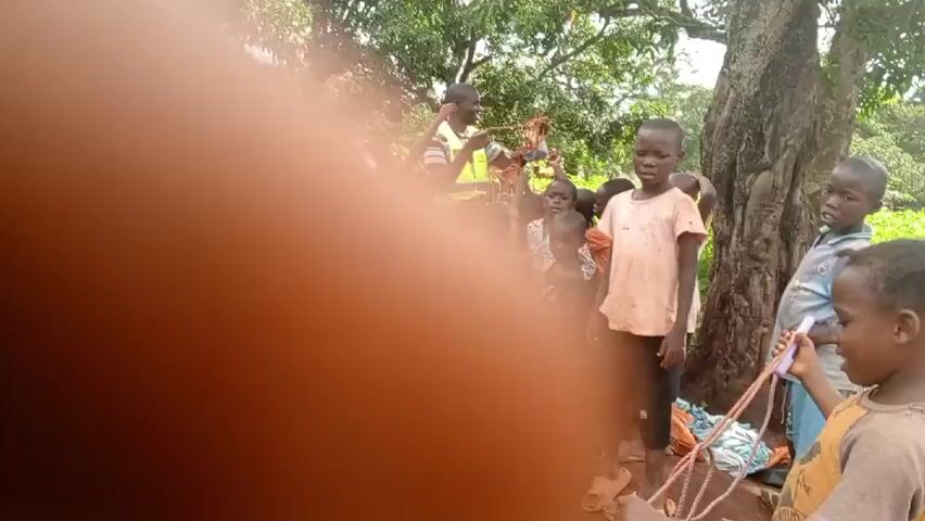 The video shows a group of children outdoors under a tree. There appears to be a person out of focus in the foreground blocking the view. The children are mostly standing and looking in different directions. Some are holding ropes. In the background, there is a building with a metal roof and lots of green vegetation. Then a man comes along and gives something to the kids who reach for it and raise their hands.