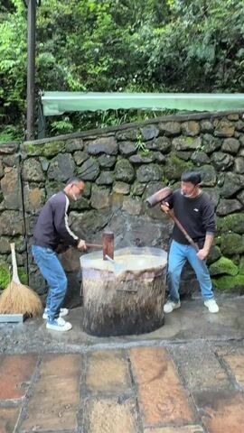 Two Asian men are pounding a thick white substance in a large wooden tub outdoors. The men wear casual clothes, face masks, and alternate striking the substance with large wooden mallets while chanting. The tub has a dark, weathered exterior. Behind the men, there's a mossy stone wall and lush green vegetation. A broom leans against the wall beside the tub. The ground appears to be wet and paved.