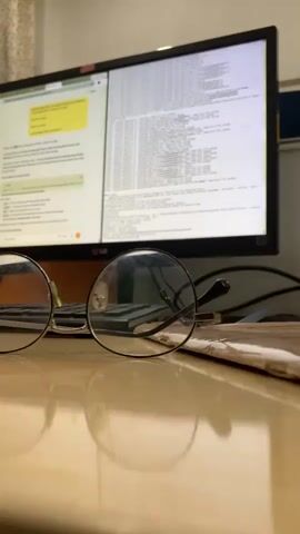 The video displays a close-up, low-angle shot of a pair of eyeglasses resting on a wooden desk. In the background, a computer monitor displays lines of text, suggesting a work or study environment. The lighting is dim, with the monitor providing the primary light source. The focus is on the eyeglasses, with the background slightly blurred. The camera appears to be static for the duration of the clip.